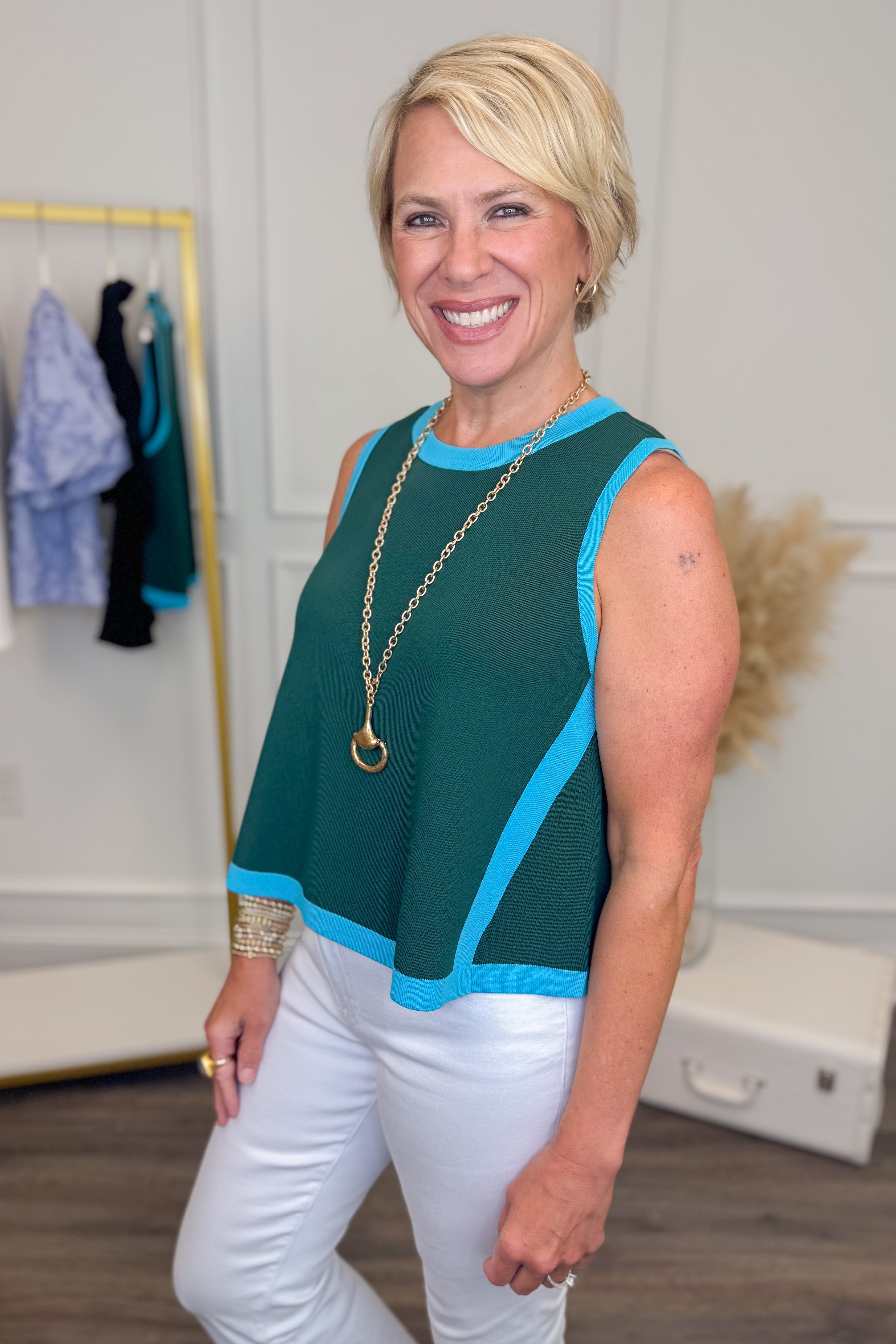 side view of woman wearing a teal sleeveless knit sweater white white pants. 
