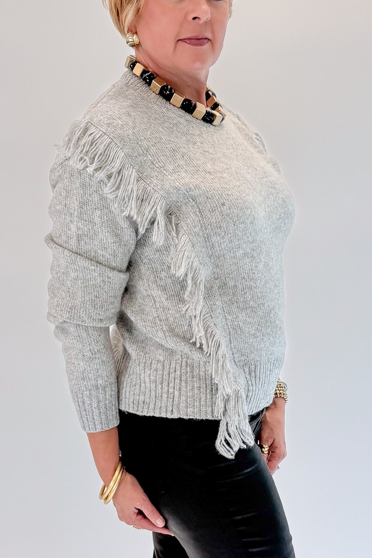 Side detail view of woman wearing Swagger Boutique’s Grey Fringe Sweater with jeans against white studio background.