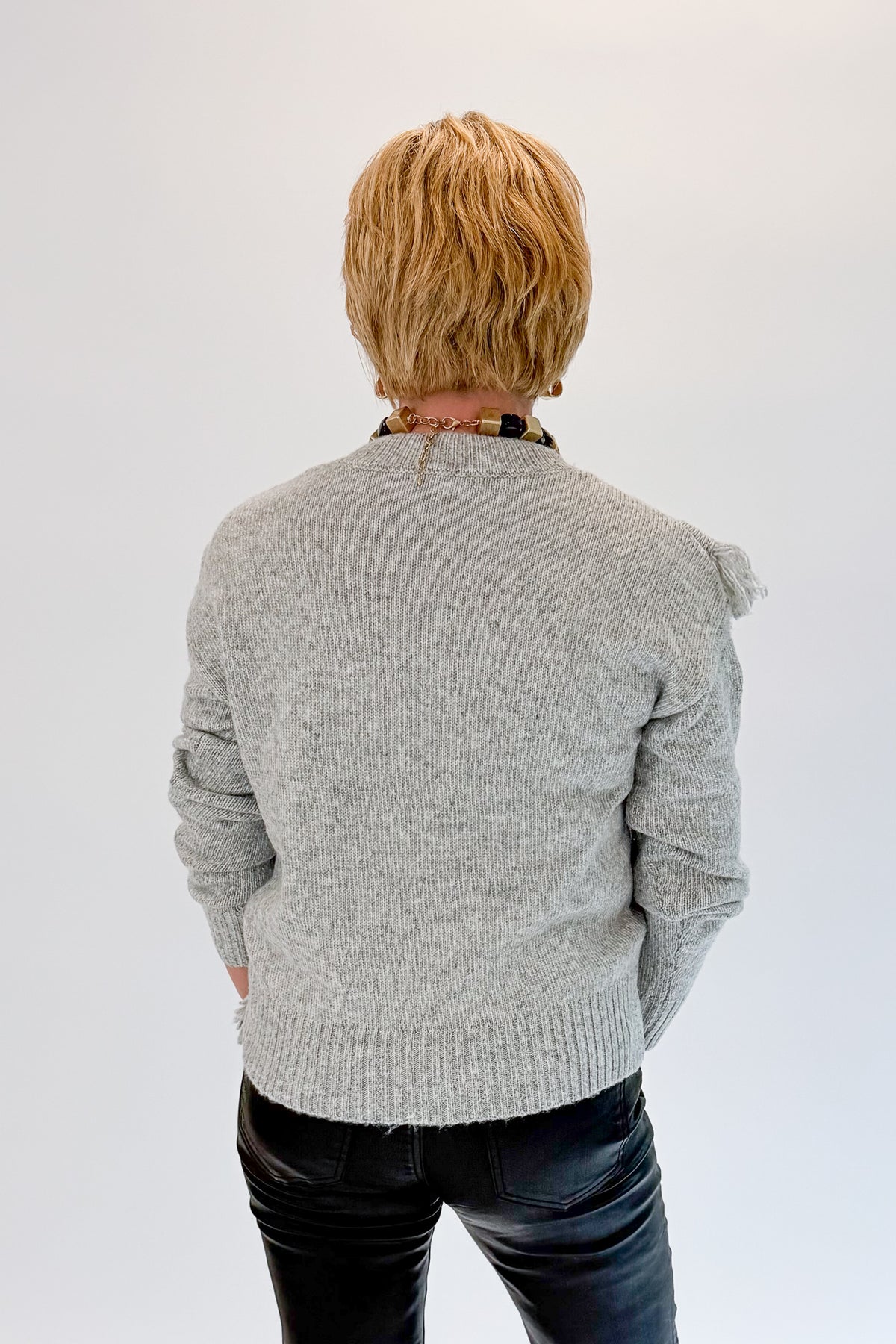 Back view of woman wearing Swagger Boutique’s Grey Fringe Sweater with jeans against white studio background.