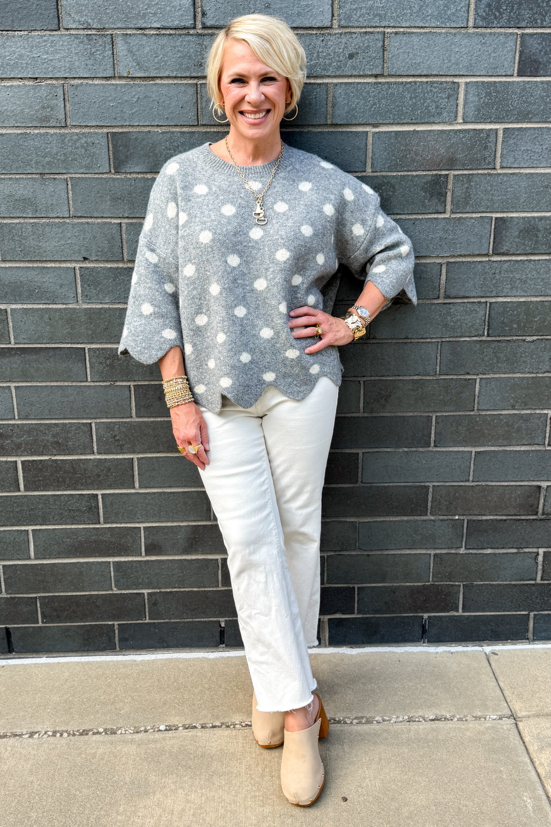 Front view of woman wearing Swagger Boutique’s Grey Polka Dot Short Sleeve Sweater by Entro with jeans standing outside against a brick wall.