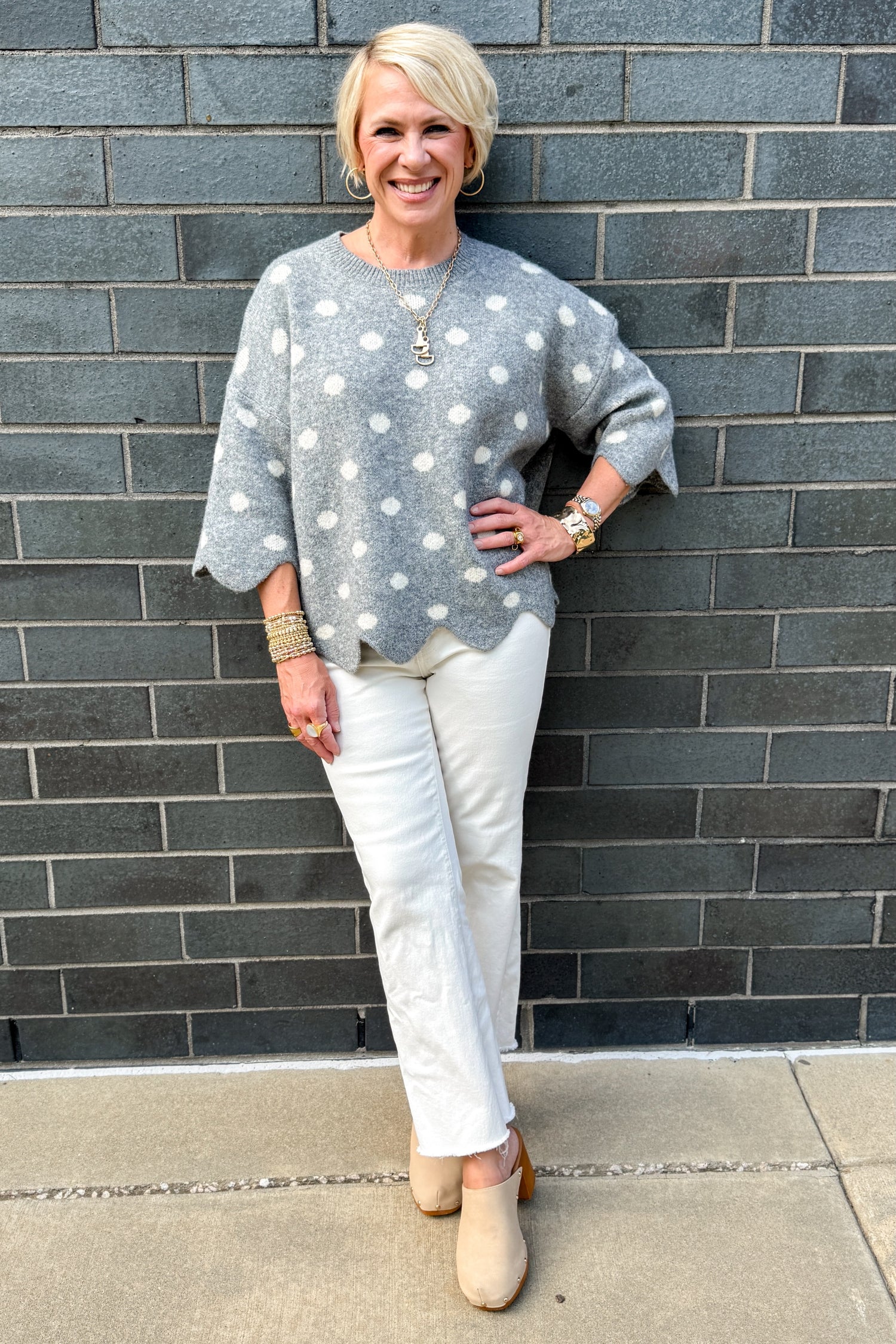 Front view of woman wearing Swagger Boutique’s Grey Polka Dot Short Sleeve Sweater by Entro with jeans standing outside against a brick wall.