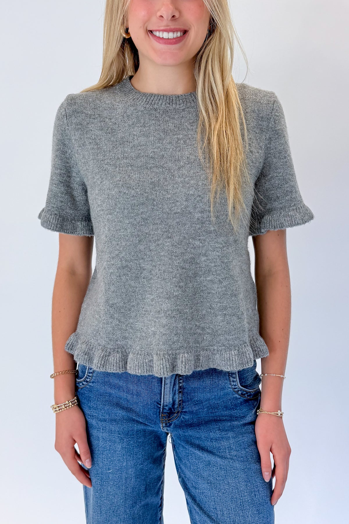 Front detail view of woman wearing Swagger Boutique’s Grey Ruffle Hem Sweater with jeans against white studio background.