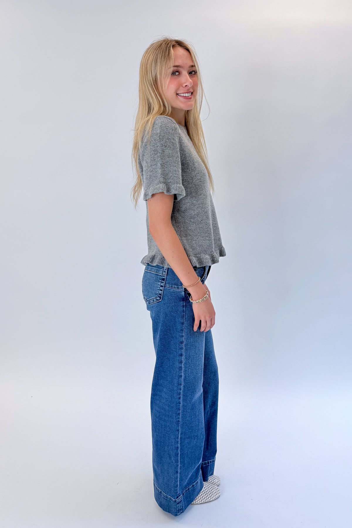 Side view of woman wearing Swagger Boutique’s Grey Ruffle Hem Sweater with jeans against white studio background.