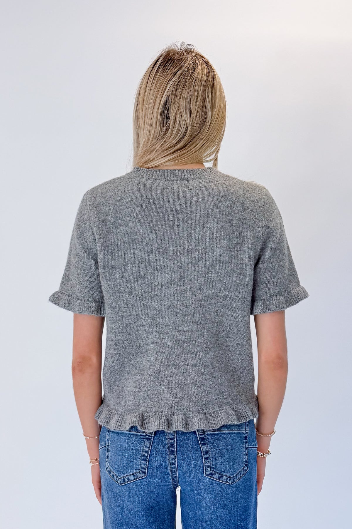 Back view of woman wearing Swagger Boutique’s Grey Ruffle Hem Sweater with jeans against white studio background.