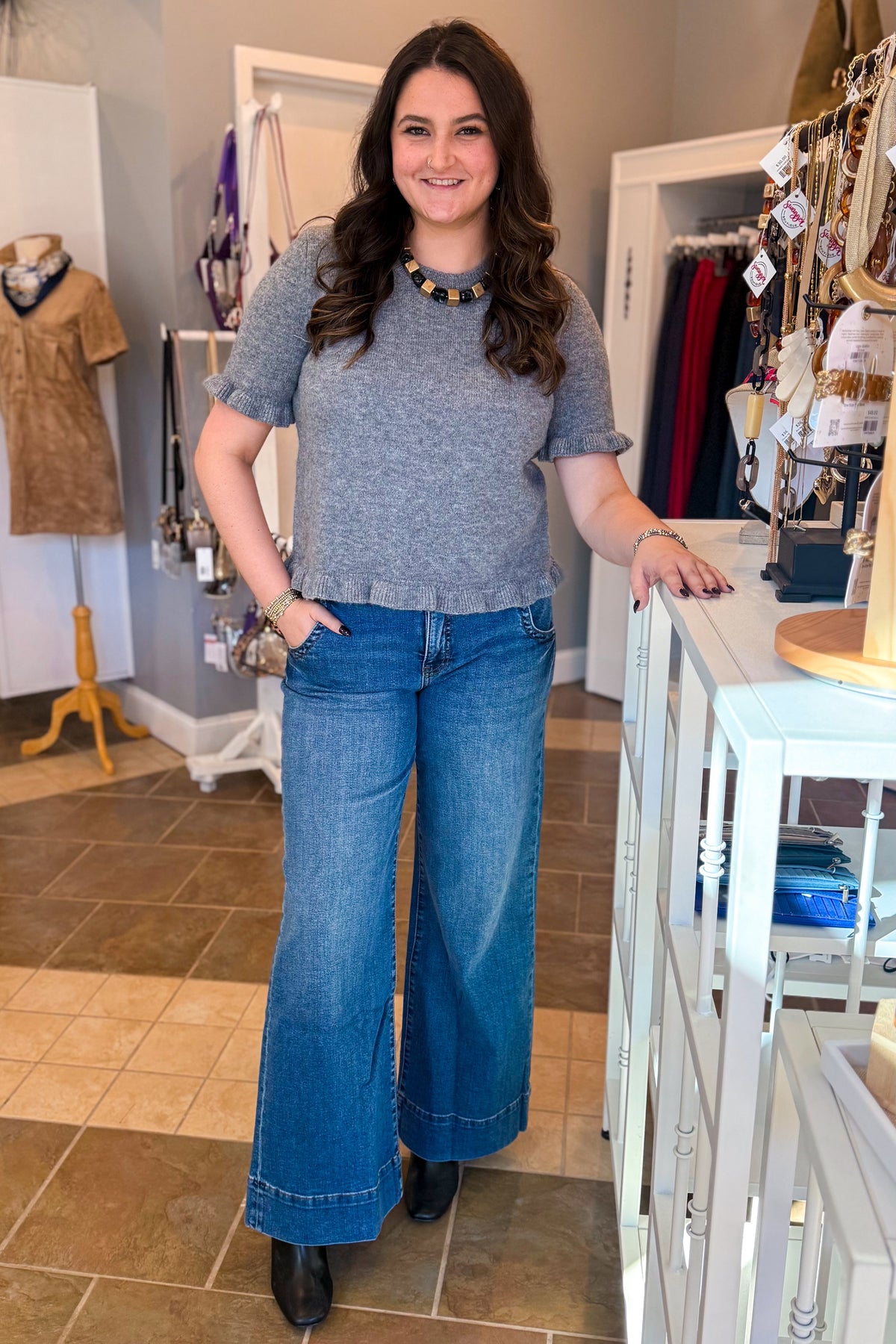 Front view of woman wearing Swagger Boutique’s Grey Ruffle Hem Sweater with jeans standing at the store.