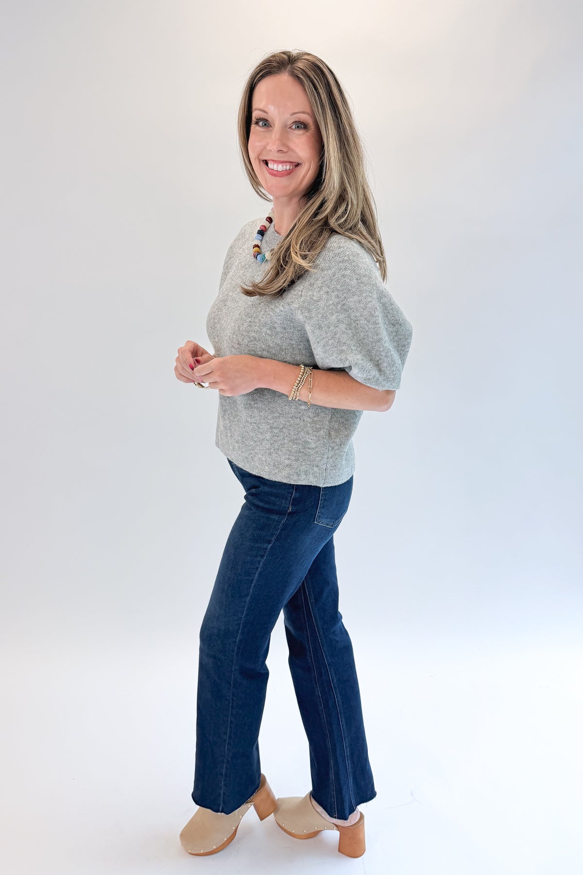 Side view of woman wearing Swagger Boutique’s Grey Short Sleeve Sweater with jeans against white studio background.