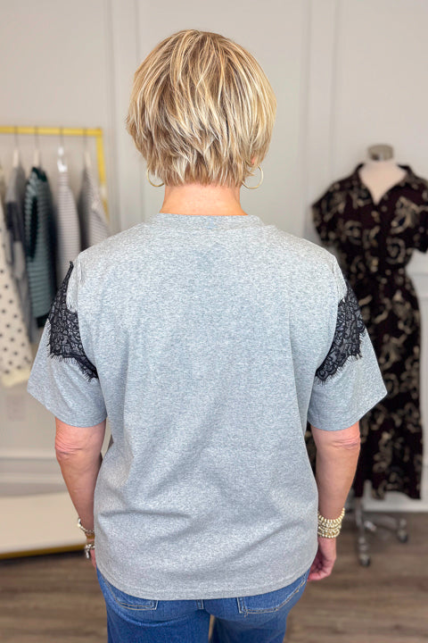 Back view of woman wearing Swagger Boutique’s Grey &amp; Black Lace Contrast Tee with jeans with clothing hanging in the background.