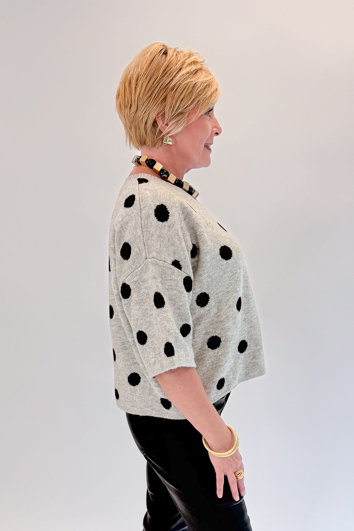 Side view of woman wearing Swagger Boutique’s Grey &amp; Black Polka Dot Short Sleeve Cardigan with jeans against white studio background.