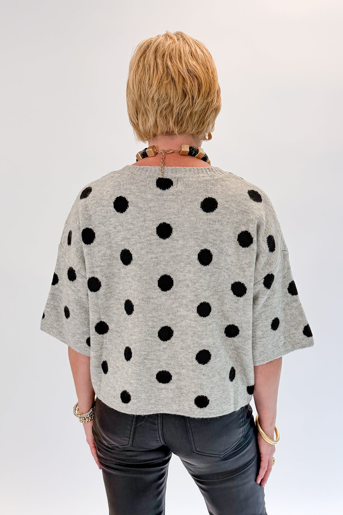 Back view of woman wearing Swagger Boutique’s Grey &amp; Black Polka Dot Short Sleeve Cardigan with jeans against white studio background.