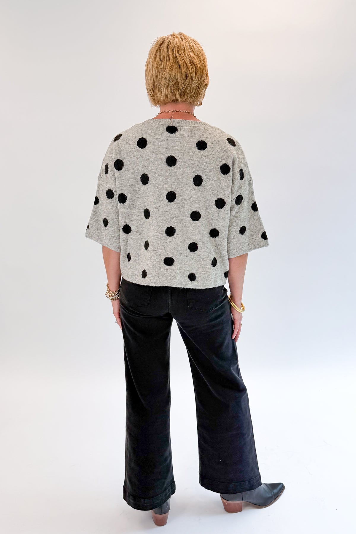 Back view of woman wearing Swagger Boutique’s Grey &amp; Black Polka Dot Short Sleeve Cardigan with jeans against white studio background.