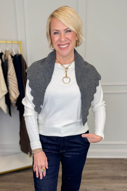 A smiling blonde woman wearing a white ribbed long-sleeve top, dark blue jeans, and a gray scalloped knit scarf, standing in a boutique setting with a clothing rack visible in the background.
