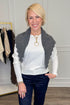 A smiling blonde woman wearing a white ribbed long-sleeve top, dark blue jeans, and a gray scalloped knit scarf, standing in a boutique setting with a clothing rack visible in the background.