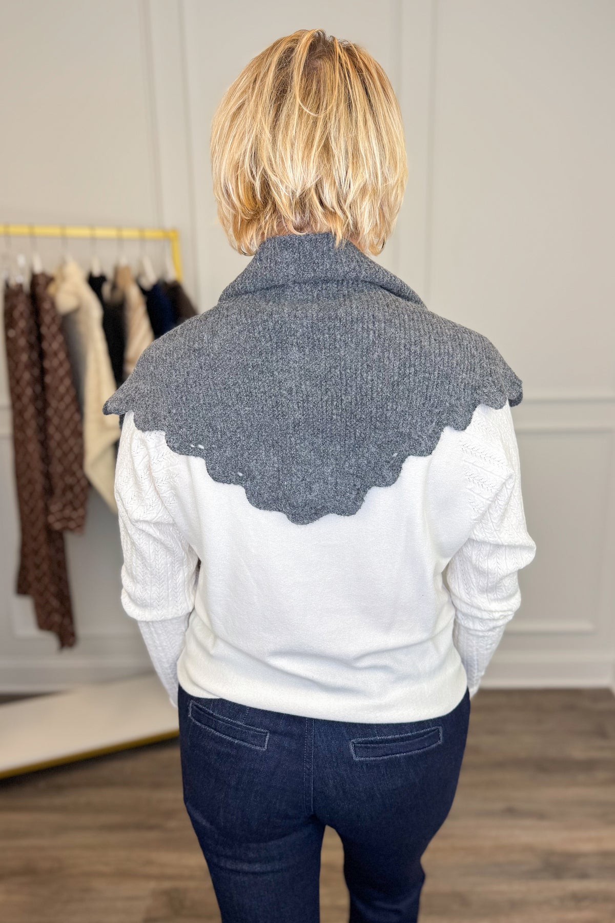 A woman viewed from behind wearing a gray knit cowl scarf with a scalloped edge over a white long-sleeve top and dark blue jeans, standing in a boutique setting.