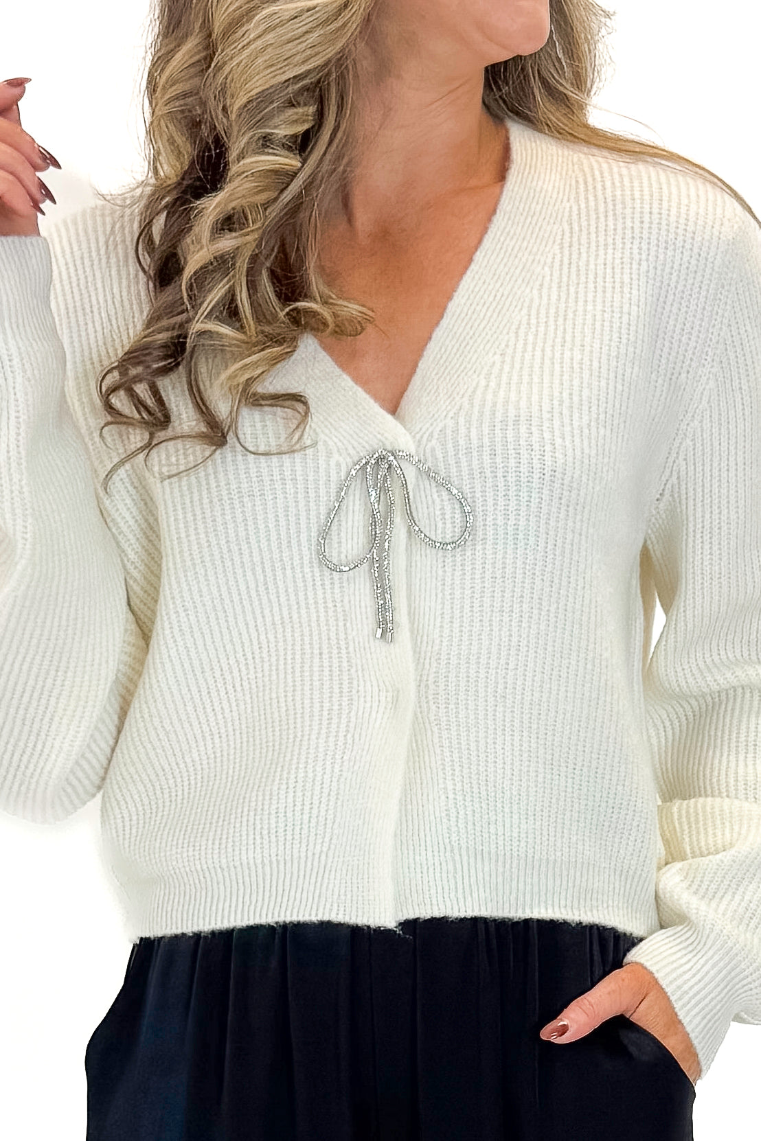 Front detail view of woman wearing Cream Cardigan With Rhinestone Bow with pants against white studio background.