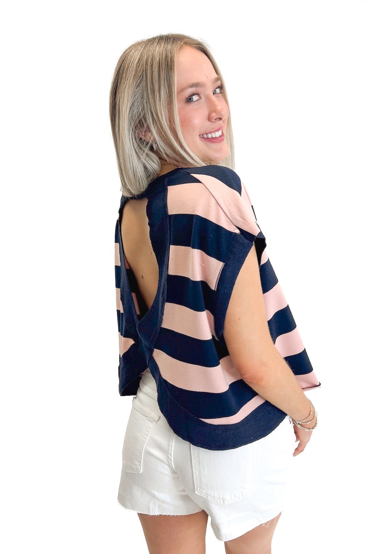 Side view of woman wearing Navy &amp; Pink Striped Cut Out Back Tee with jean shorts against white studio background.