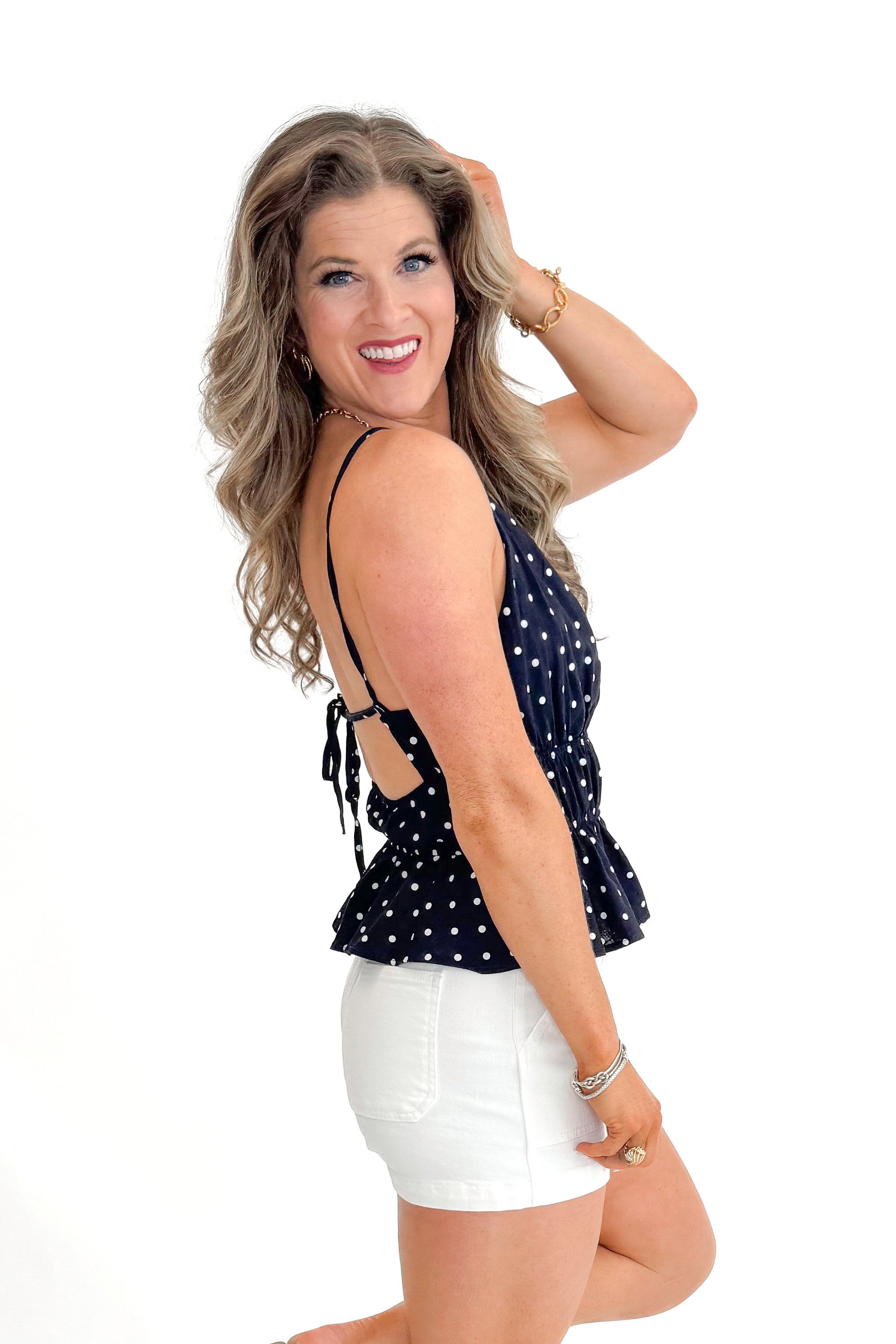 Side view of woman wearing Navy Polka Dot Peplum Tank with jean shorts against white studio background.