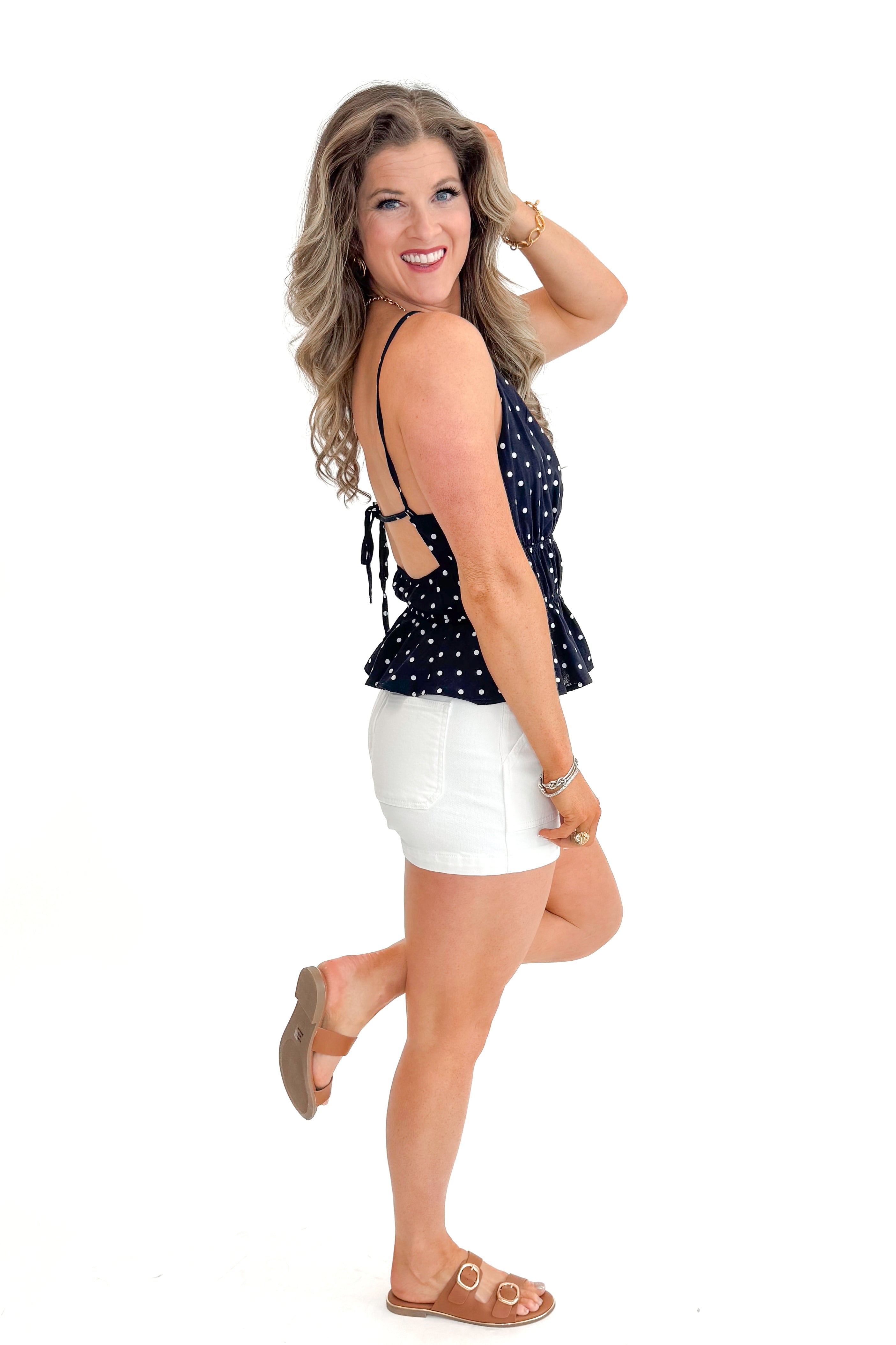 Side view of woman wearing Navy Polka Dot Peplum Tank with jean shorts against white studio background.