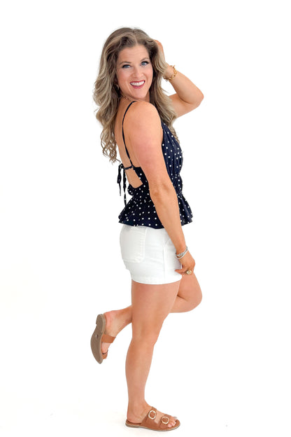 Side view of woman wearing Navy Polka Dot Peplum Tank with jean shorts against white studio background.