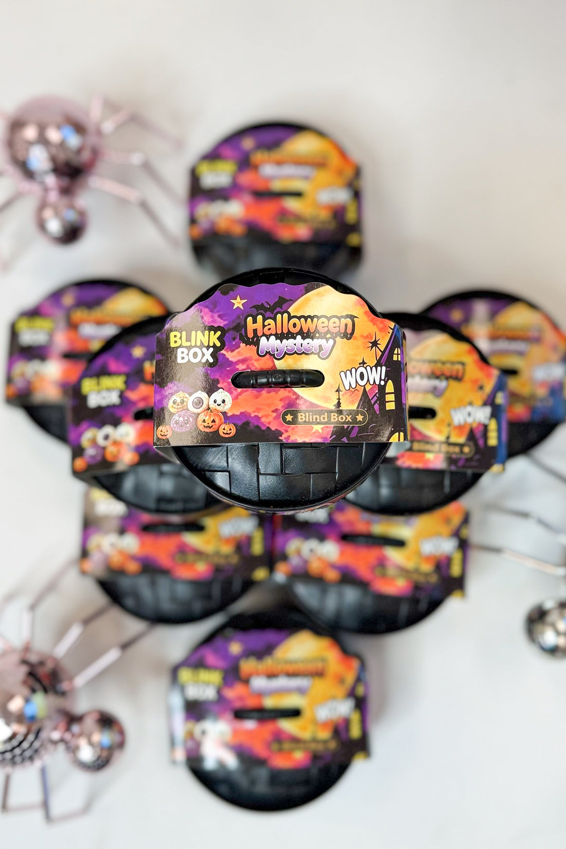 Several Halloween-themed blind box packages arranged in a circle on a white surface, with spider decorations in the corners. The front package is most prominent and shows the text &