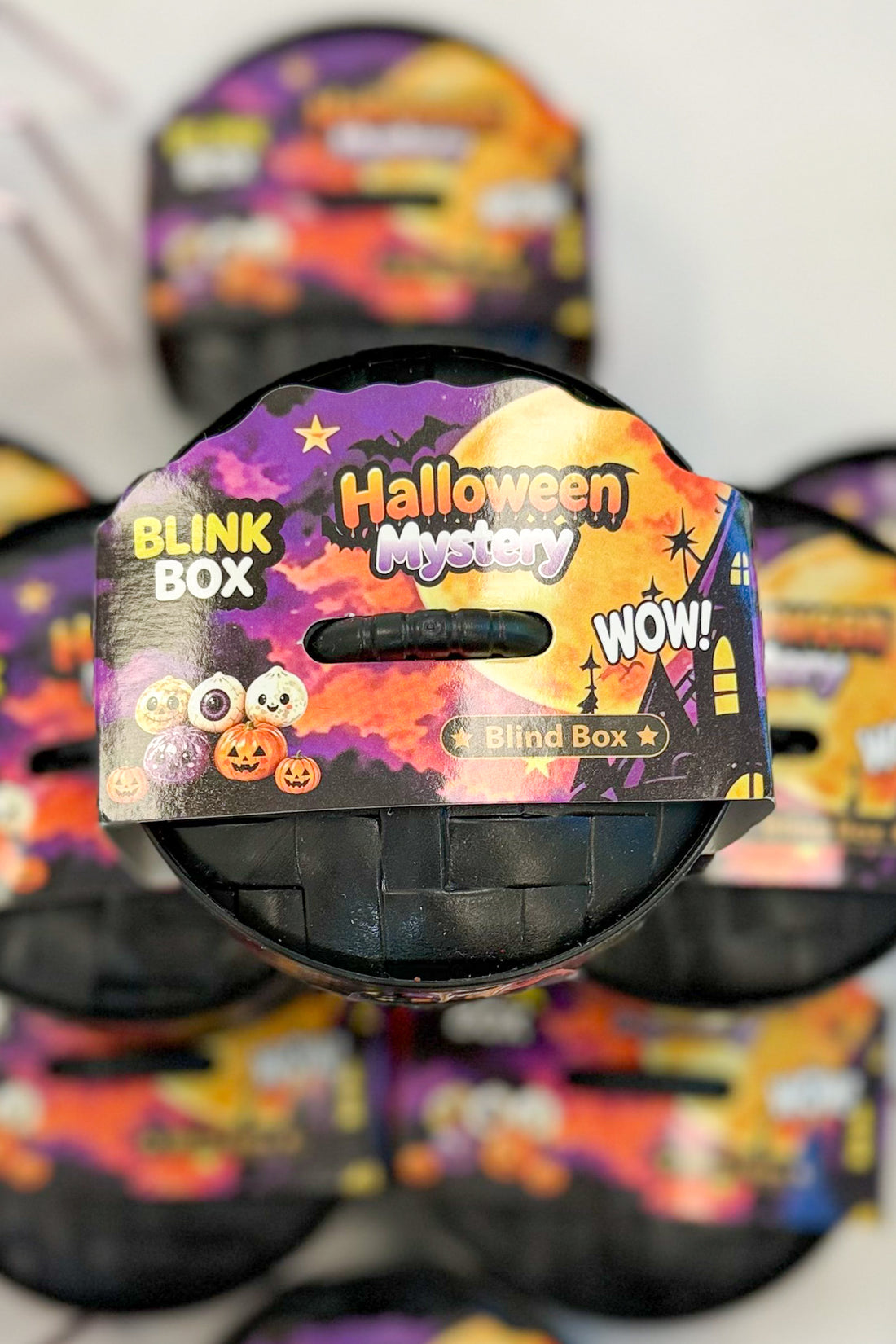 A Halloween mystery blind box toy with a black plastic container and a colorful Halloween-themed cardboard lid. The lid features festive graphics including pumpkins, ghosts, a full moon, and text reading &