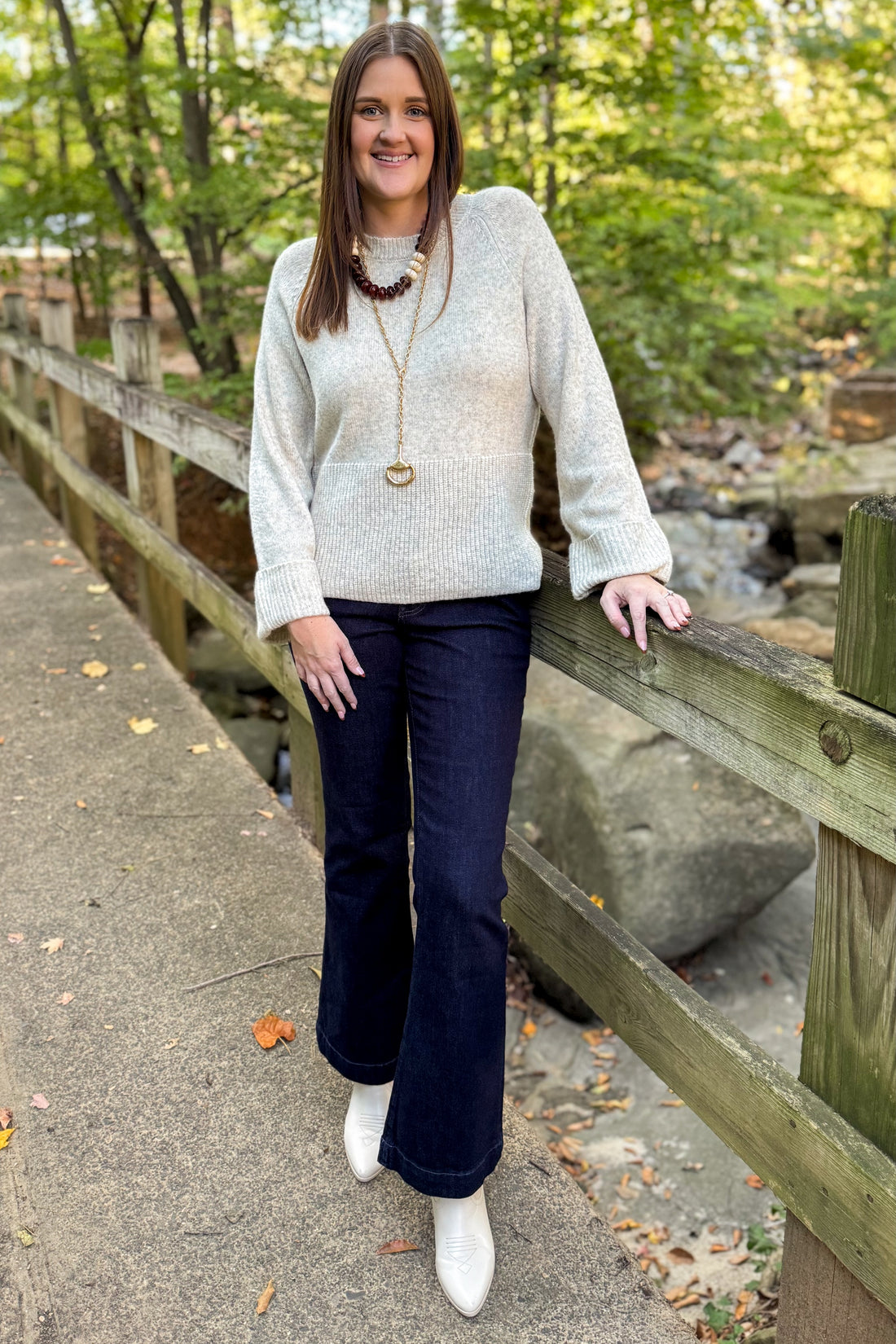 Front view of woman wearing Swagger Boutique’s Heathered Oatmeal Cuffed Sleeve Sweater with jeans standing on a bridge outside.