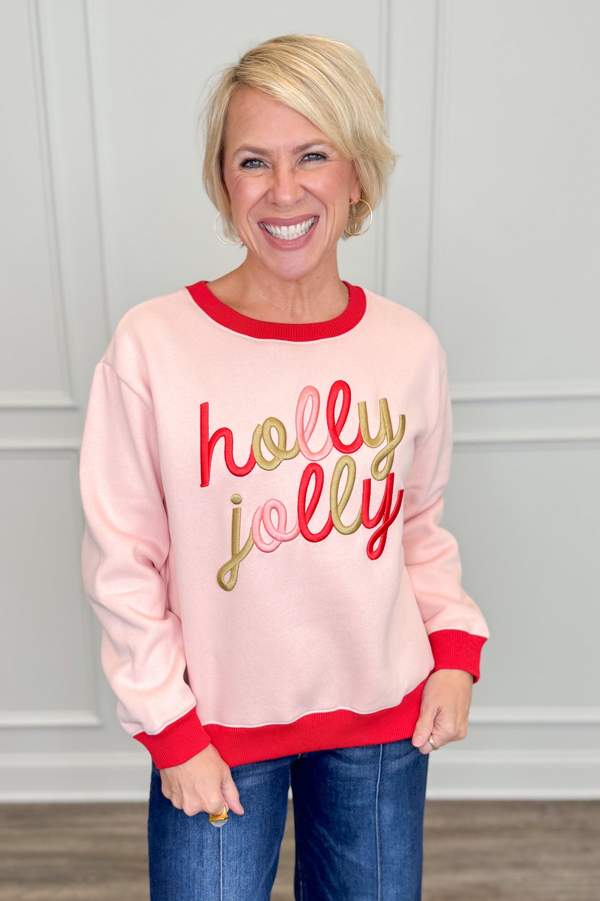 A smiling blonde woman wearing a light pink crewneck sweatshirt with red ribbed trim and &