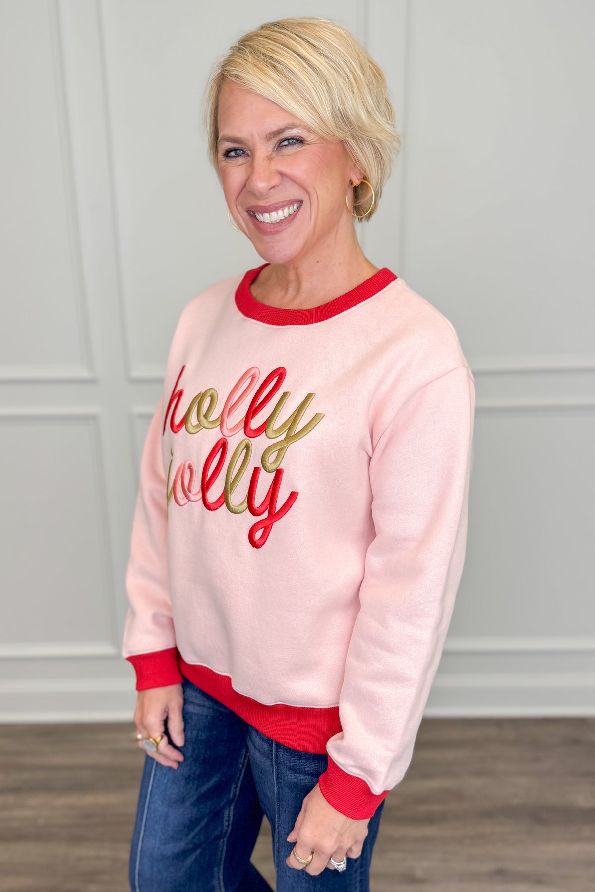 A smiling blonde woman wearing a light pink crewneck sweatshirt with red ribbed trim and &