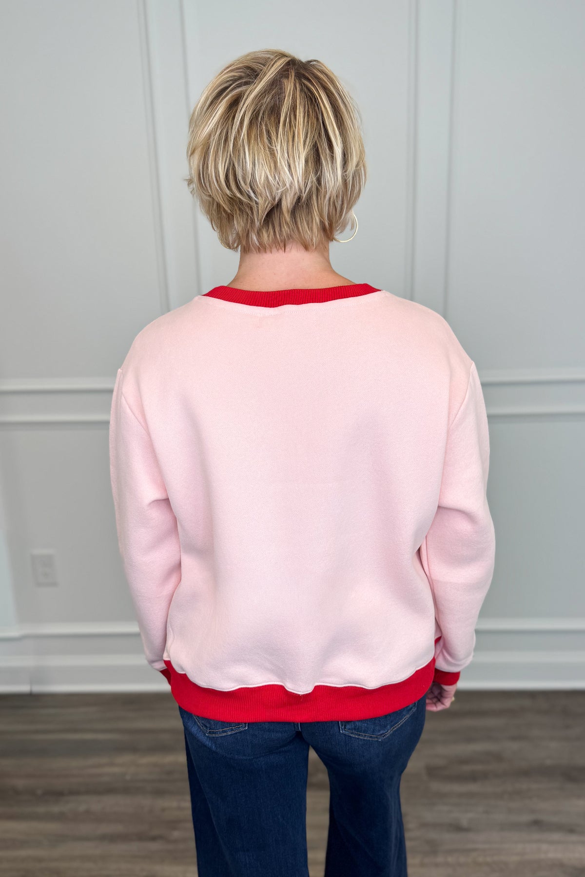 A person viewed from behind wearing a light pink crewneck sweatshirt with contrasting red trim at the collar, cuffs, and hem, paired with dark blue jeans in an indoor room with white paneled walls and wood flooring.