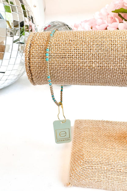Necklace with turquoise beads on a beige stand with a tag, against a neutral background.