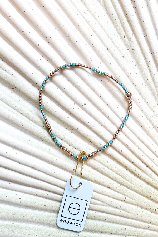 Necklace with turquoise beads and a gold clasp on a textured beige background, featuring the brand &