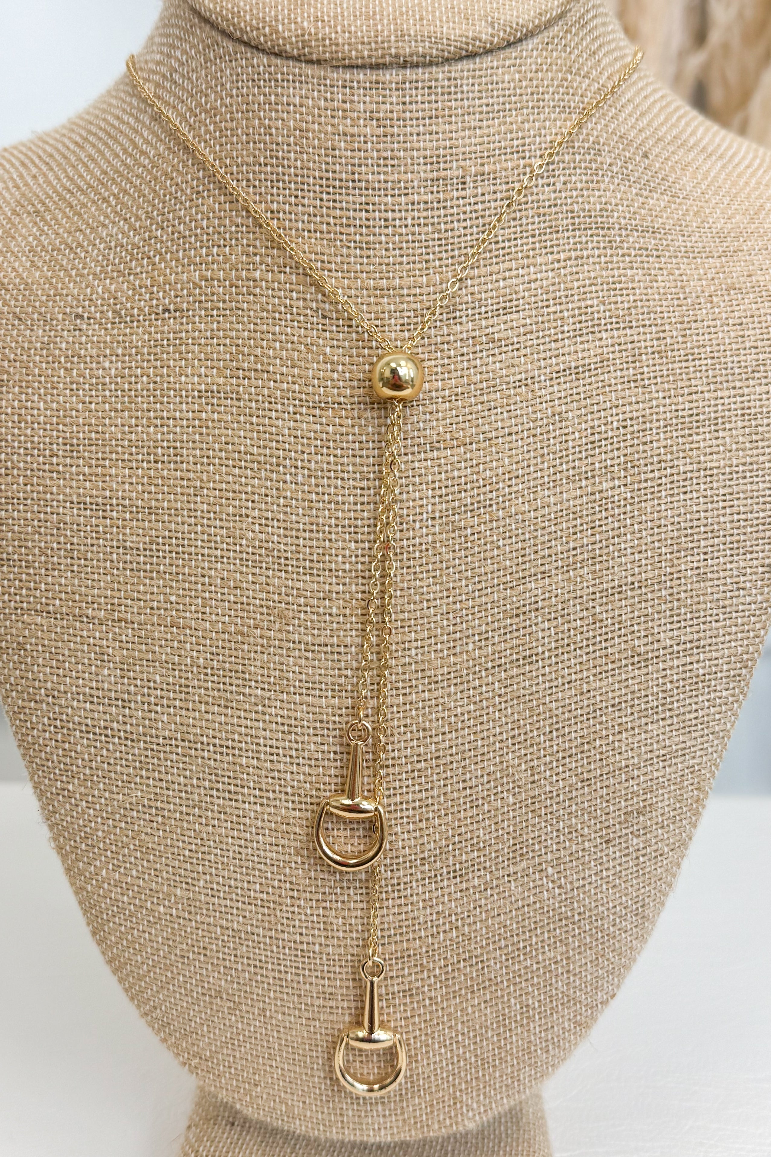 Gold-tone bolo necklace featuring a fine, reflective chain held together by a smooth, polished gold sphere slider, with delicate chain strands dropping vertically down the center each with a small horseshoe pendant