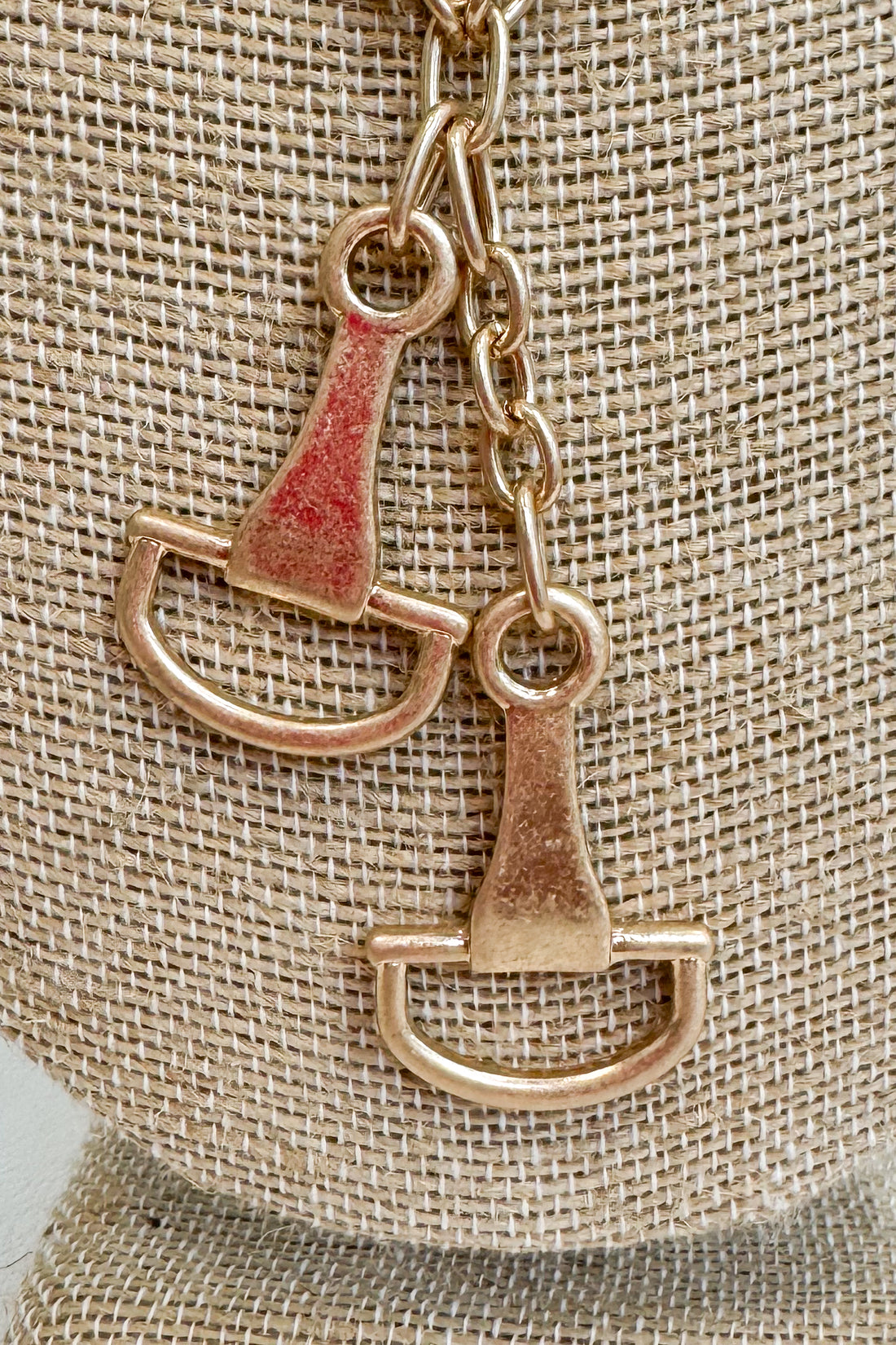 Close up of gold-tone delicate chain necklace featuring two horsebit pendants, shown on a beige jewelry bust