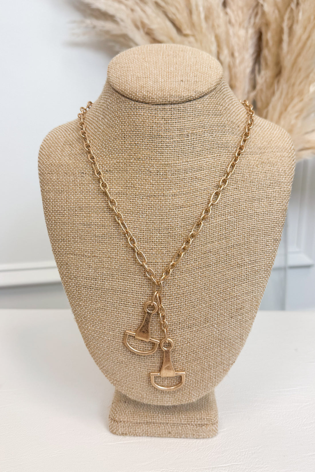 Gold-tone delicate chain necklace featuring two horsebit pendants, shown on a beige jewelry bust