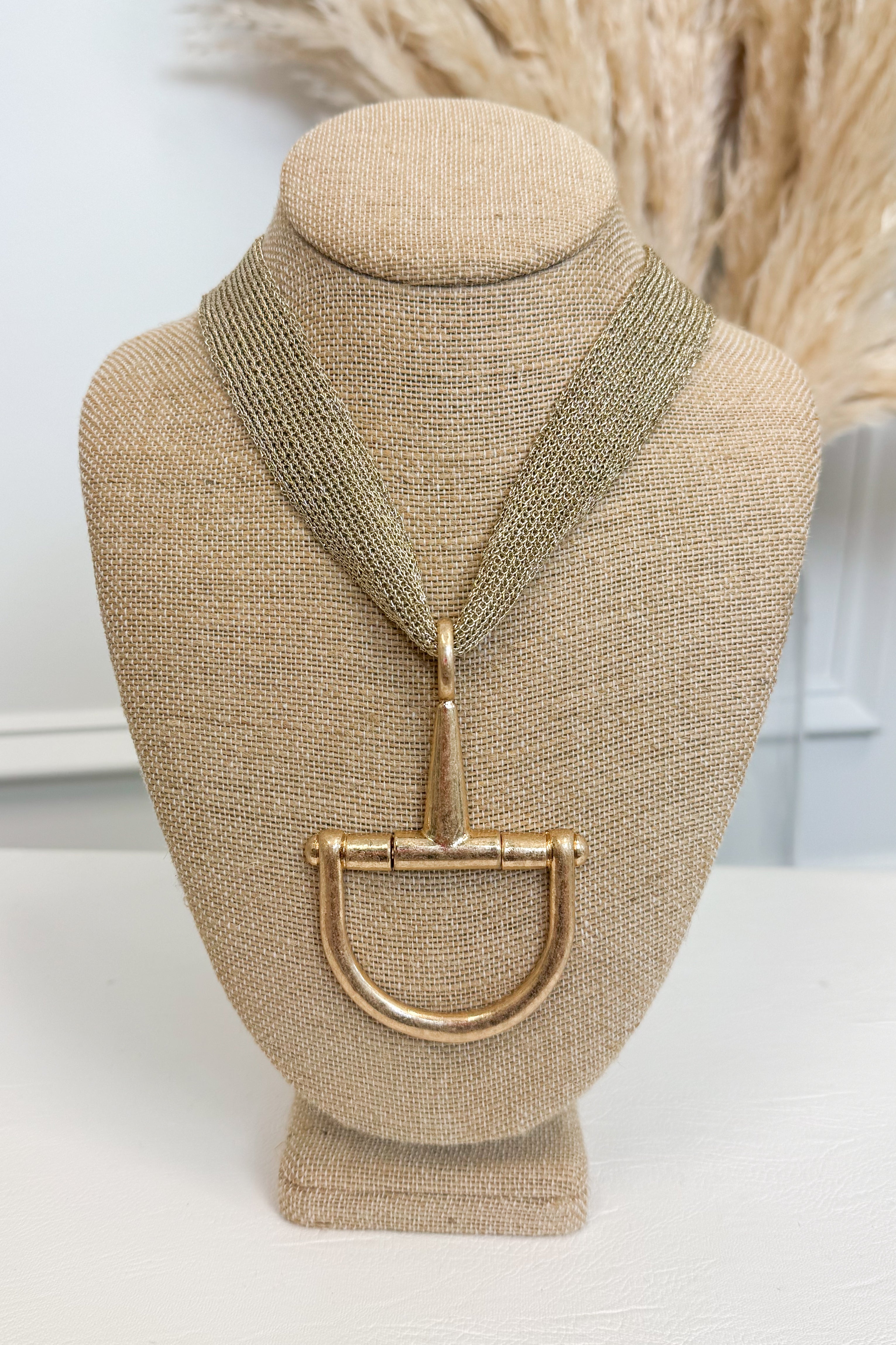 Gold-tone mesh necklace featuring a large horsebit pendant, displayed on a beige jewelry bust against a white studio background