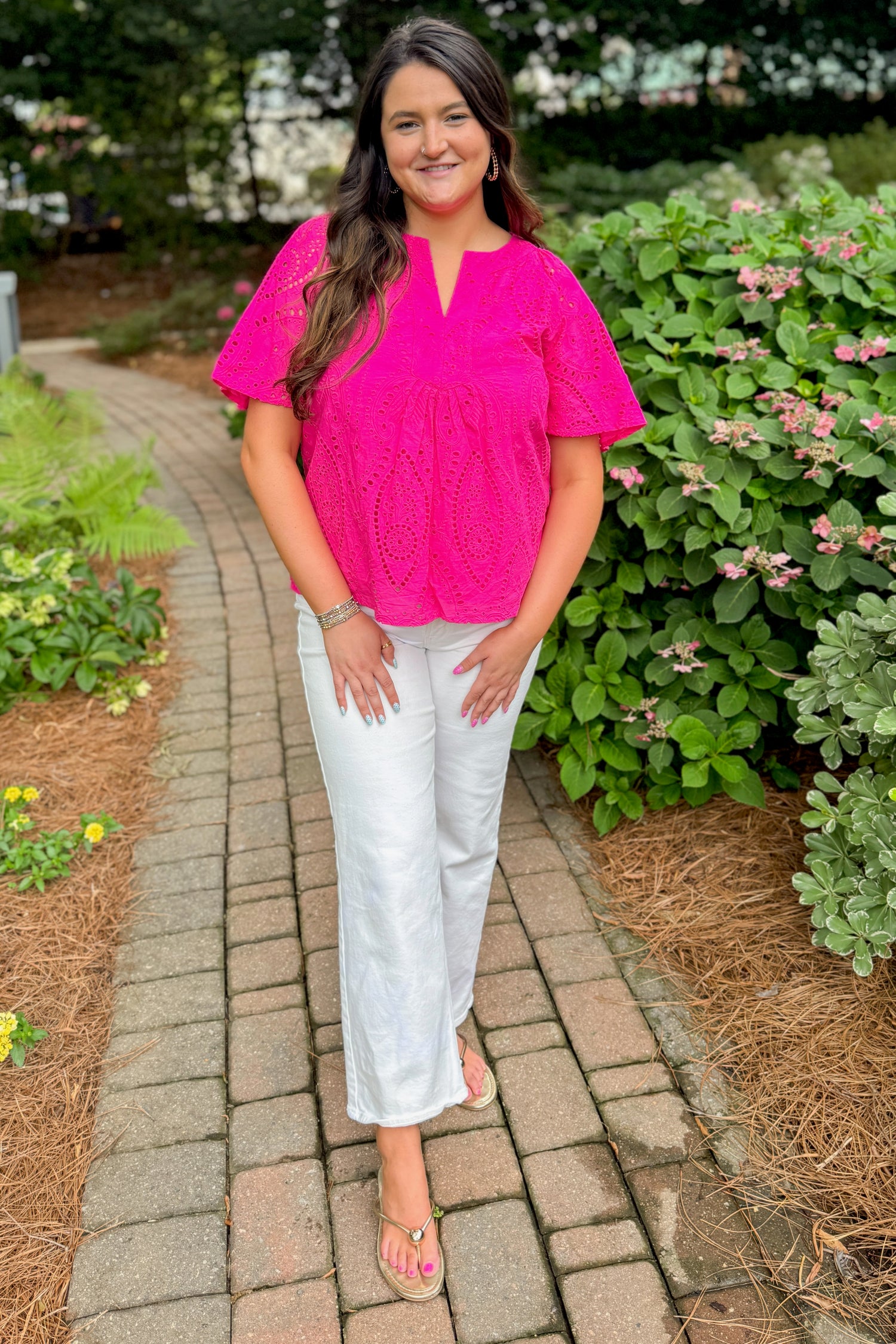 Front view of woman wearing Swagger Boutique’s Hot Pink Flutter Sleeve Eyelet Top with jeans walking down a path outside.