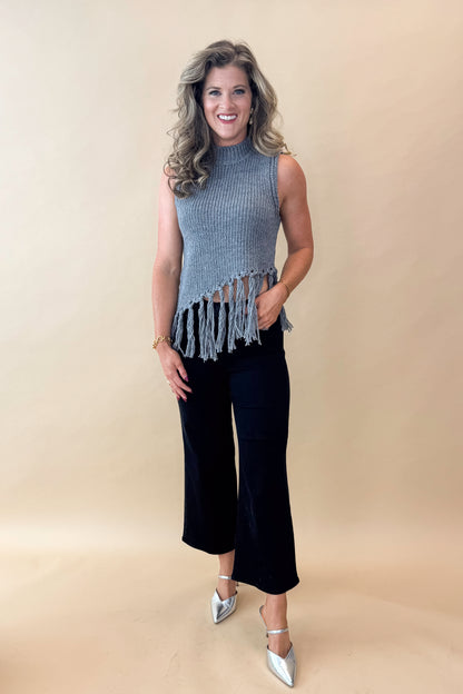 Front view of woman wearing Grey Mock Neck Fringe Detail Sleeveless Sweater and jeans against tan studio background.