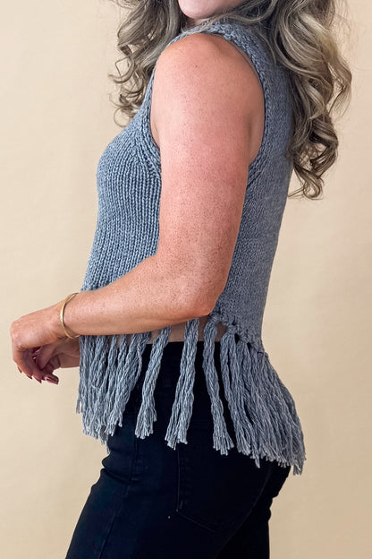 Side view of woman wearing Grey Mock Neck Fringe Detail Sleeveless Sweater and jeans against tan studio background.