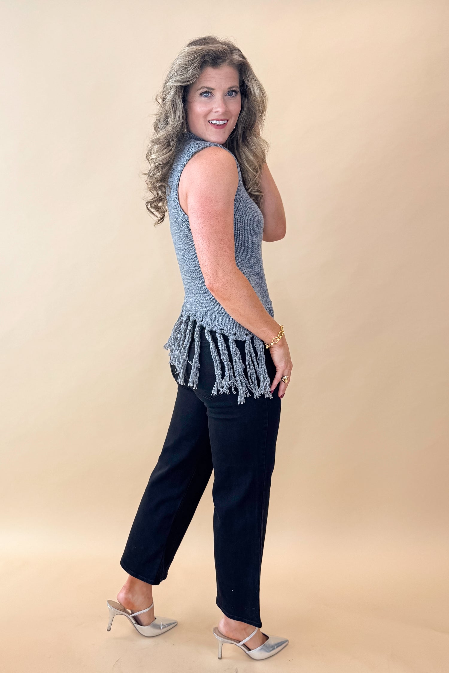 Side view of woman wearing Grey Mock Neck Fringe Detail Sleeveless Sweater and jeans against tan studio background.