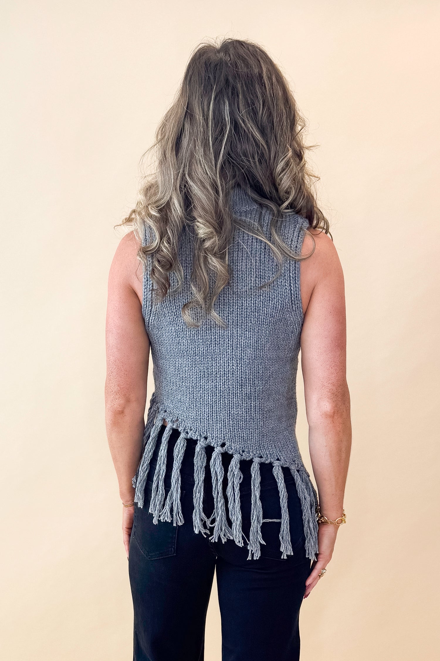 Back view of woman wearing Grey Mock Neck Fringe Detail Sleeveless Sweater and jeans against tan studio background.