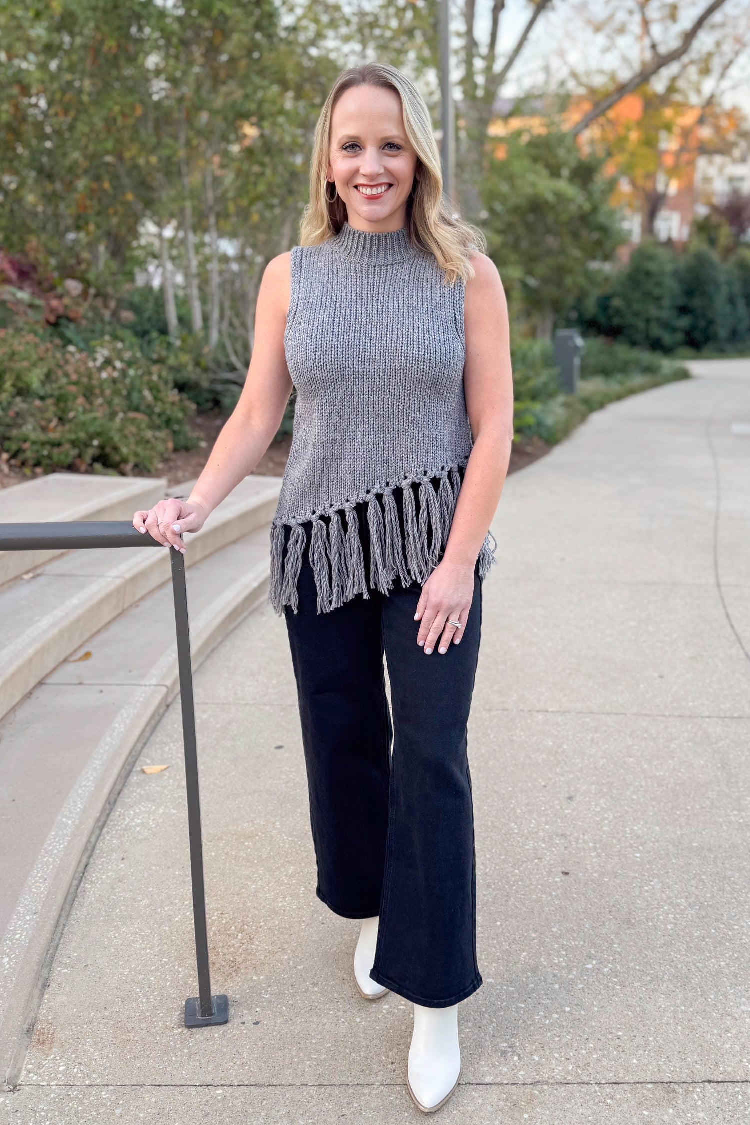 Front view of woman wearing Grey Mock Neck Fringe Detail Sleeveless Sweater and jeans outside.