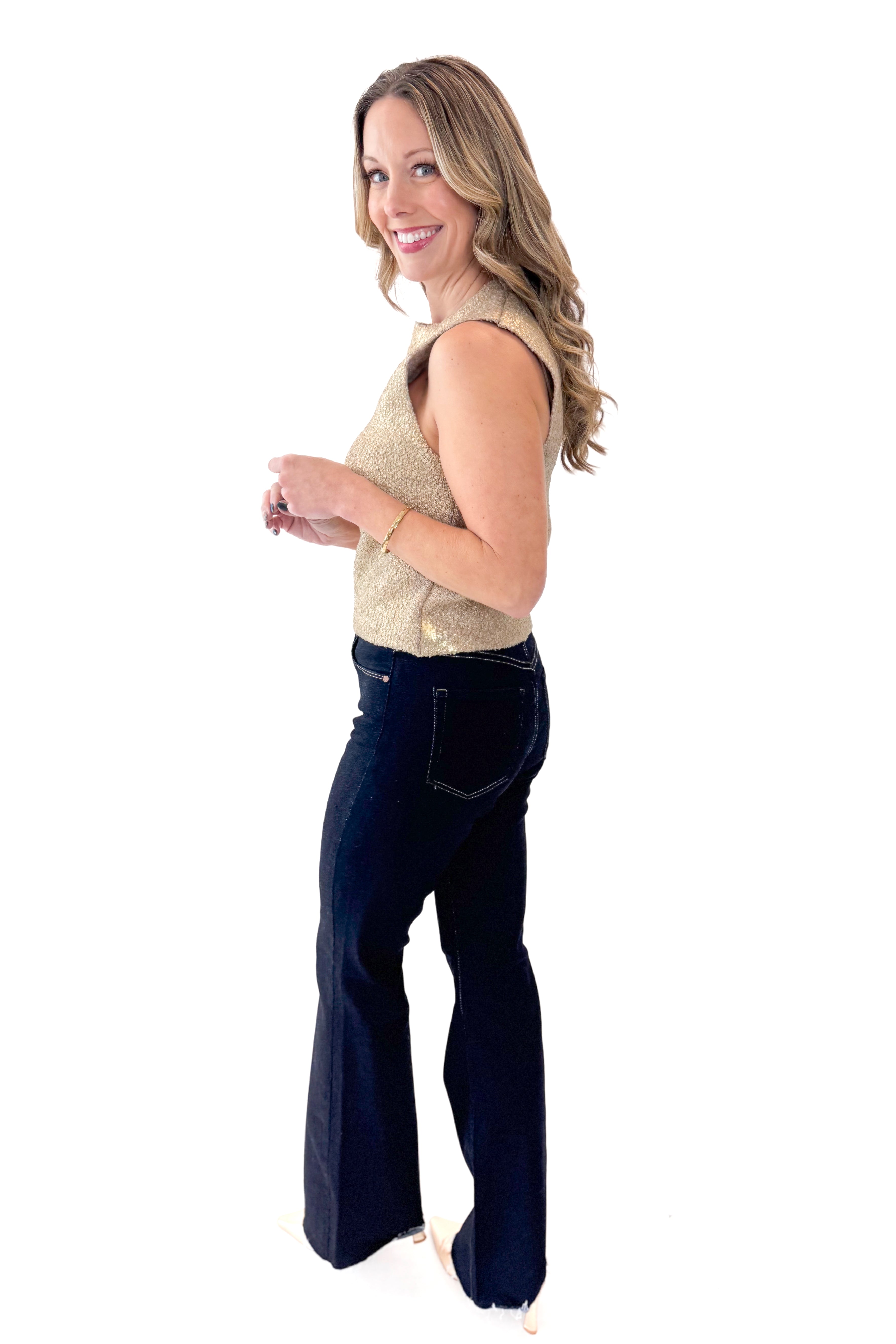 Side view of woman wearing Gold Sleeveless Sweater with jeans against white studio background.