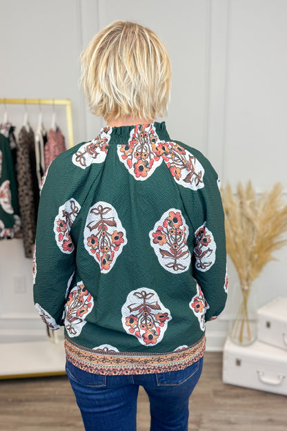 Back view of Woman wearing Hunter Green Printed &amp; Textured Puff Sleeve Top and jeans with clothes in background.