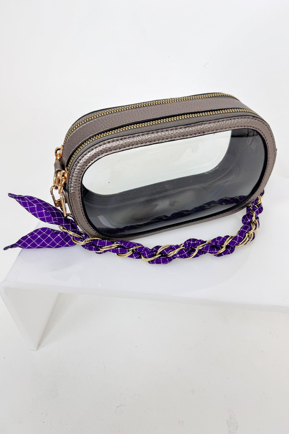 Transparent clutch with gold accents and a purple strap on a white background