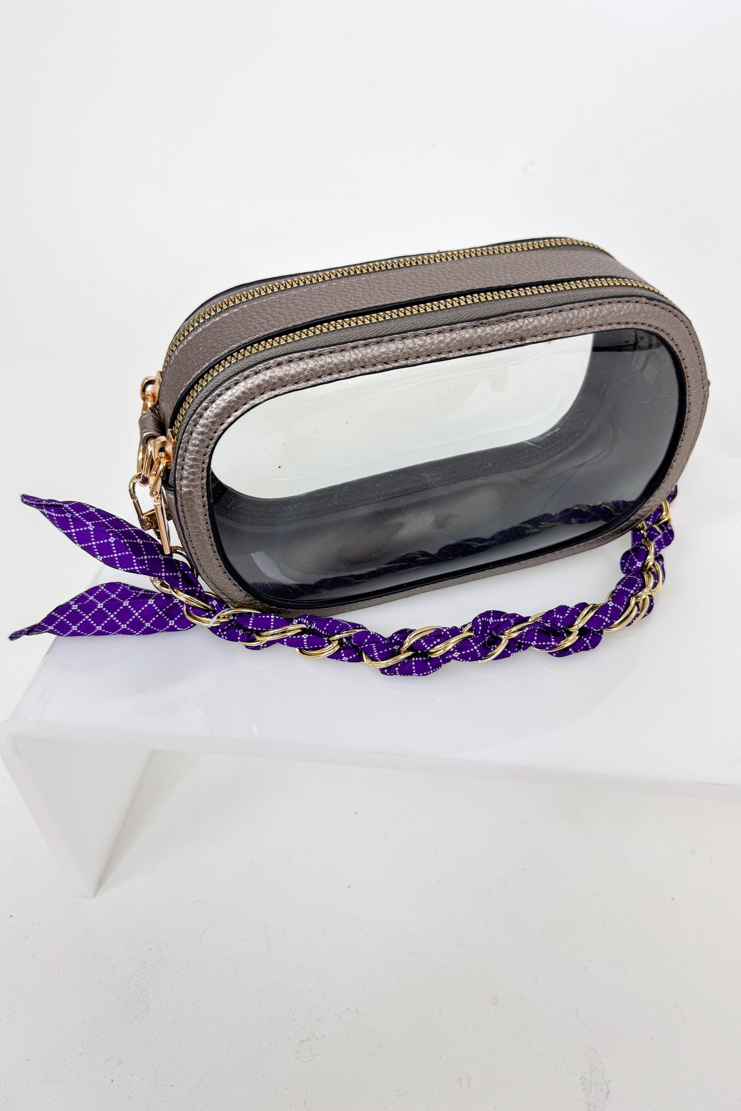 Transparent clutch with gold accents and a purple strap on a white background