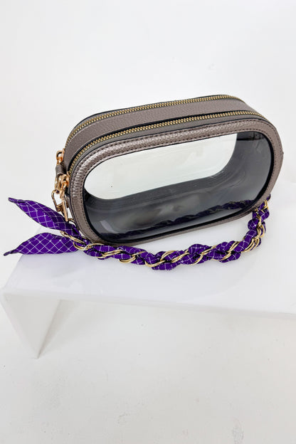 Transparent clutch with gold accents and a purple strap on a white background
