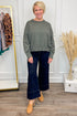 Woman wearing a green sweater and dark blue jeans standing in a room with a coat rack and decorative items.
