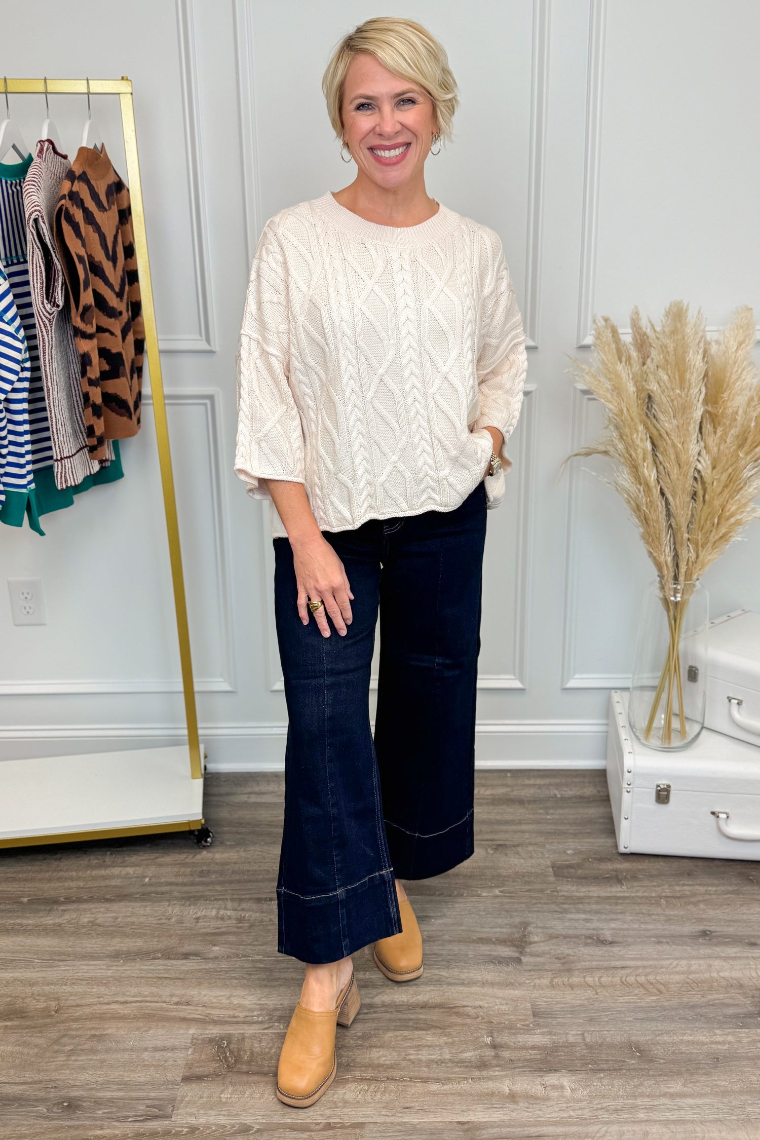Woman in a cream blouse and dark jeans standing in a room with a clothing rack and decor.
