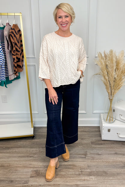 Woman in a cream blouse and dark jeans standing in a room with a clothing rack and decor.