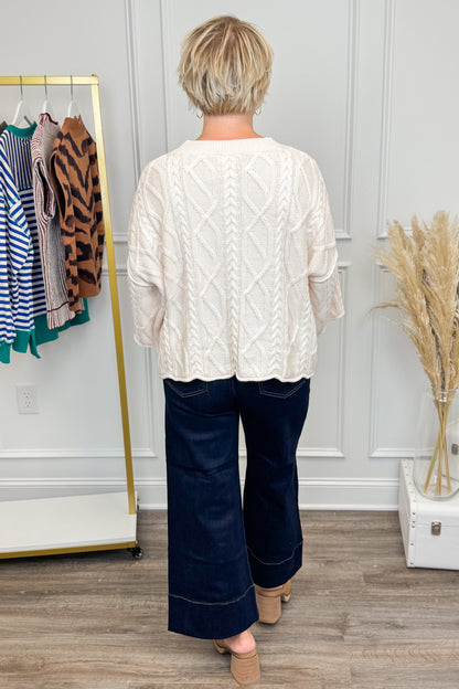 Person wearing a white cable knit sweater and navy pants in a room with a clothing rack and decorative elements.