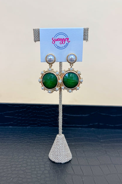 Gold earrings with green gemstones on a branded stand.